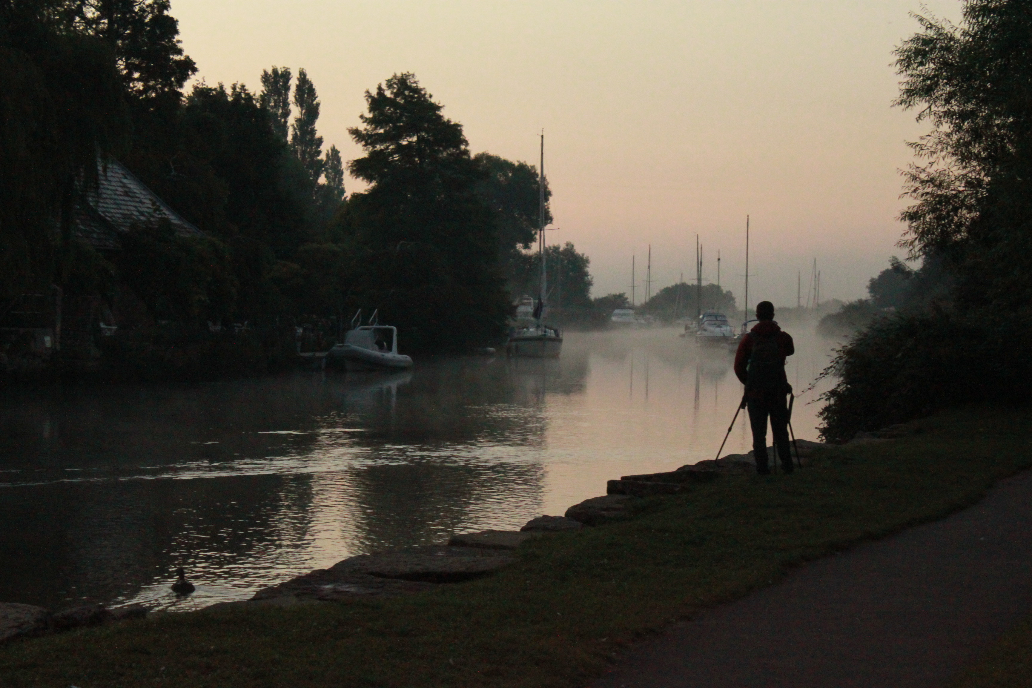 A landscape picture of a misty river scene with a siloutte of a photographer standing at the waters edge with a camera and tri pod.