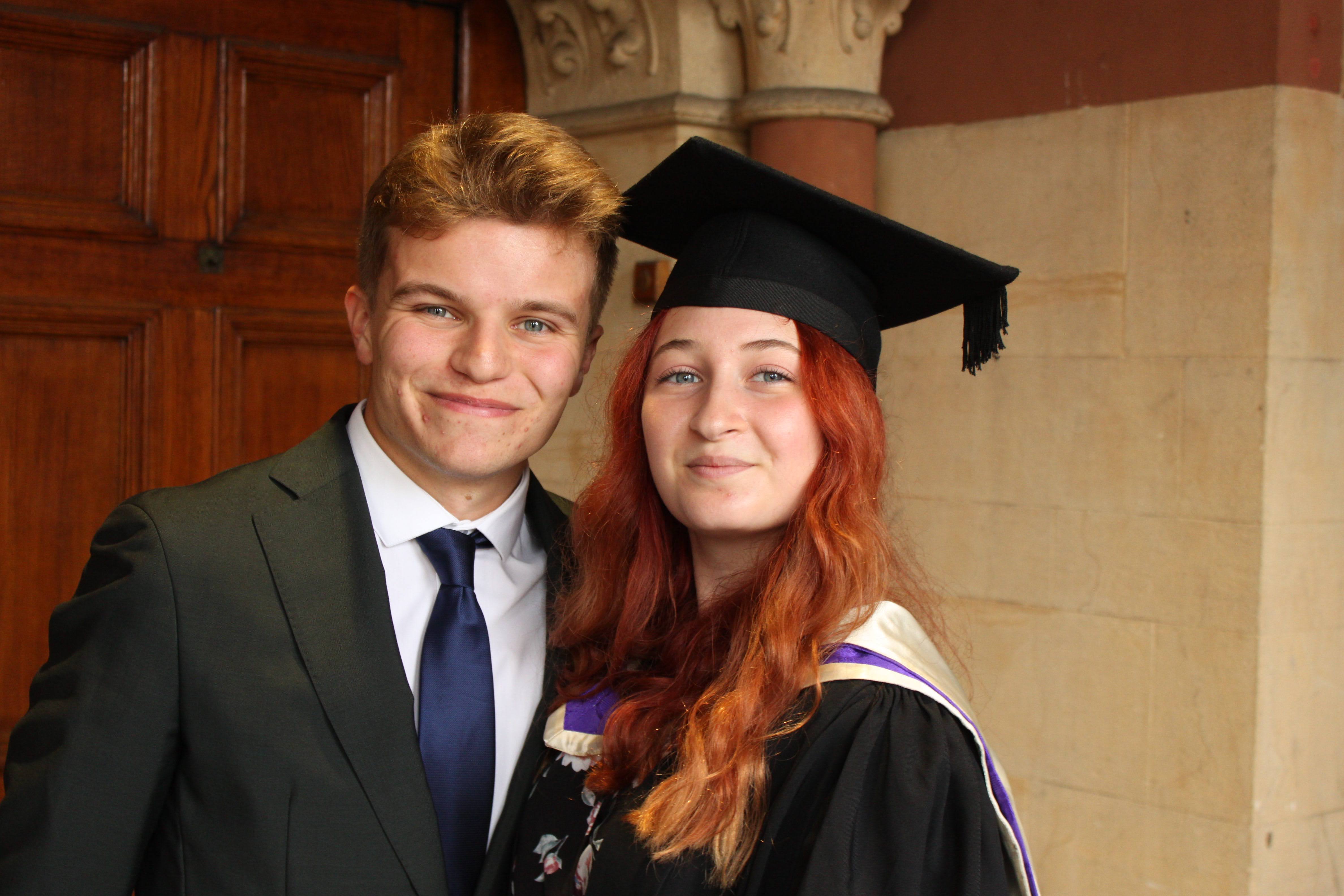 A picture of a white straight couple. Rob, a white tanned male with blonde hair who is wearing a suit, is standing on the left of Gracey, a white female with long red curly hair who is wearing a graduation gown and cap over the top of a floral dress. They are both smiling and looking at the camera.