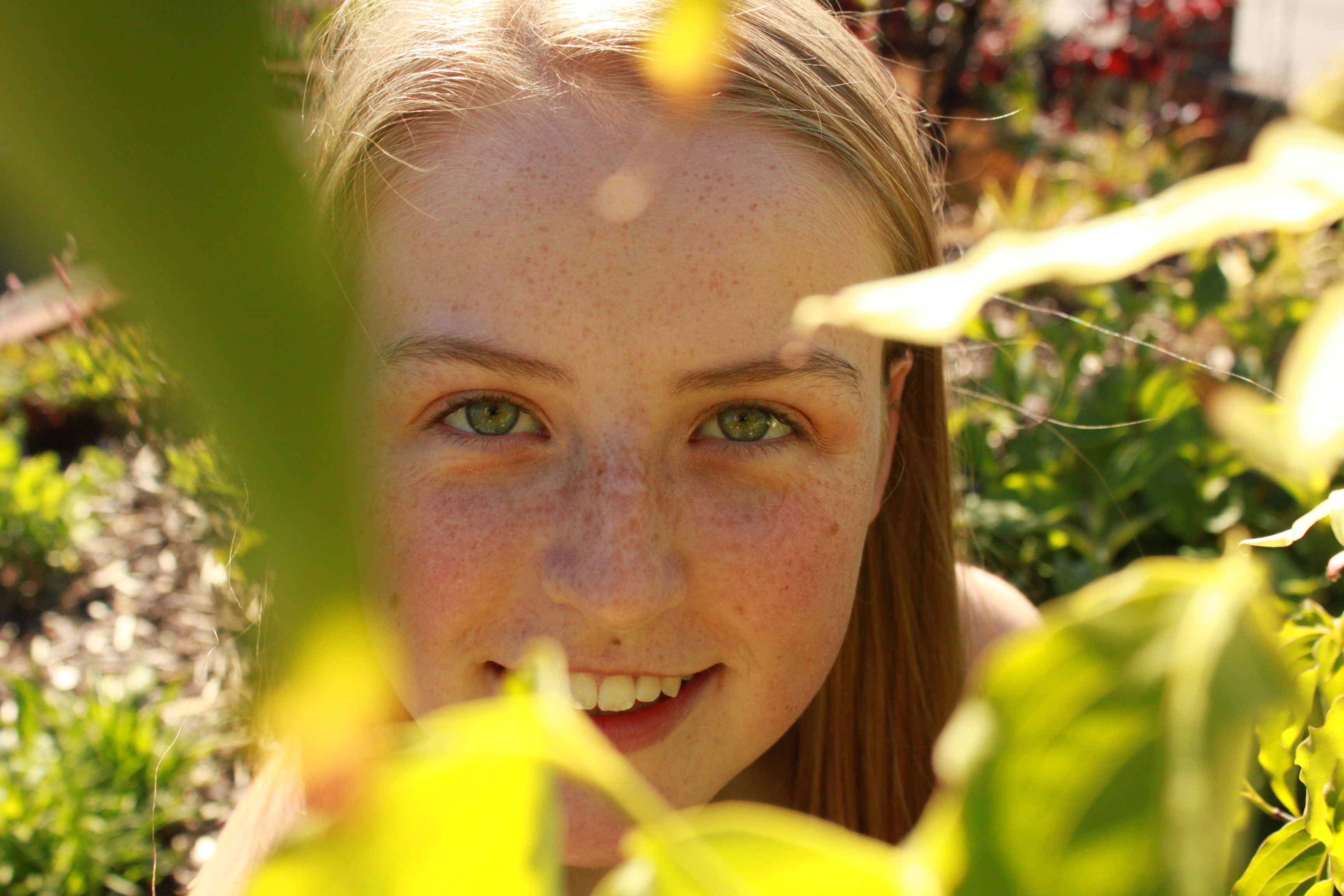 an image of a white teenager with freckles who is looking up into the camera with green leaves framing her face, highlighting her green eyes.