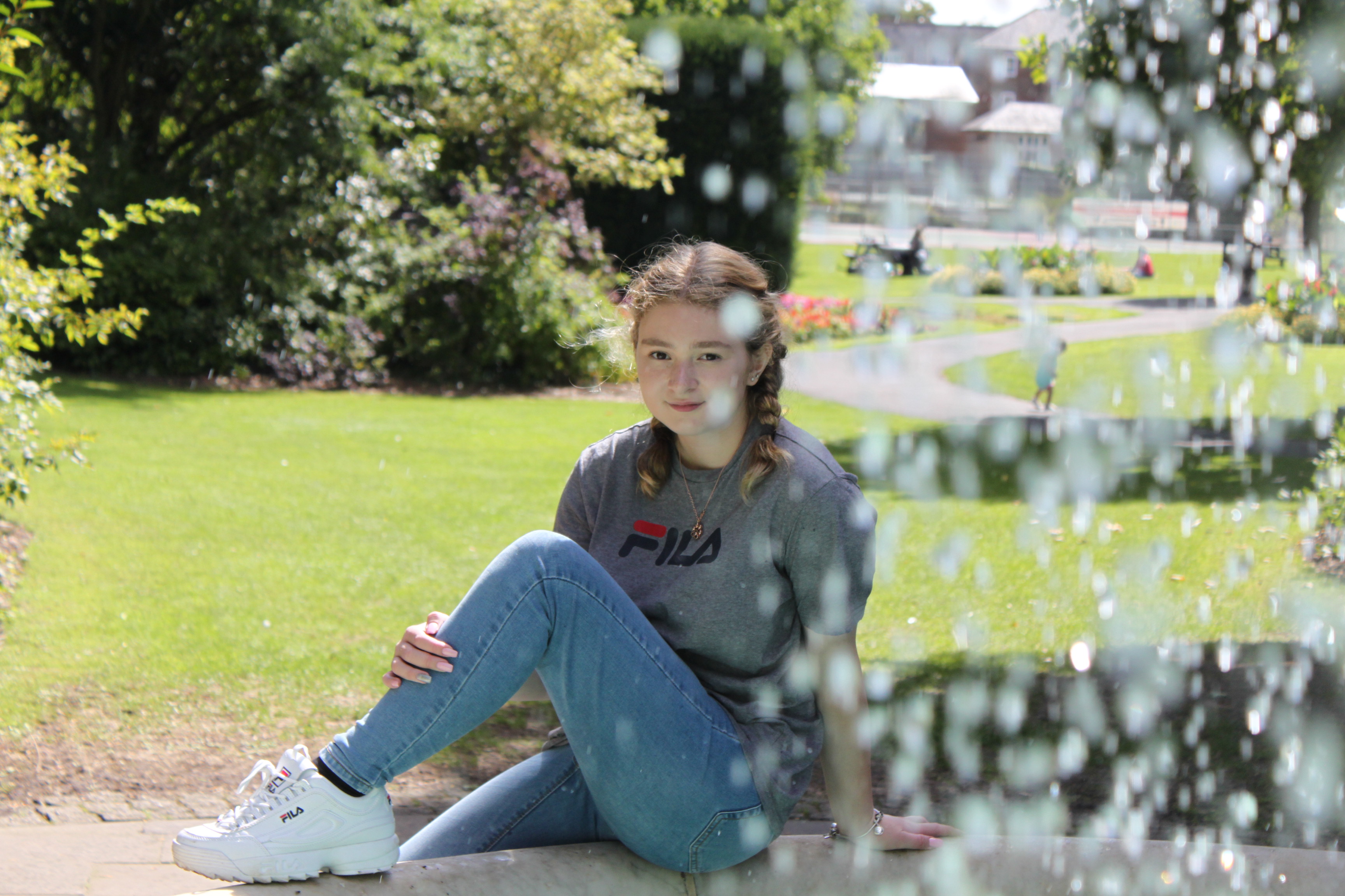 an image of a white female sitting on the ledge of a waterfountain. Infront of her is waterdroplets from the fountain.