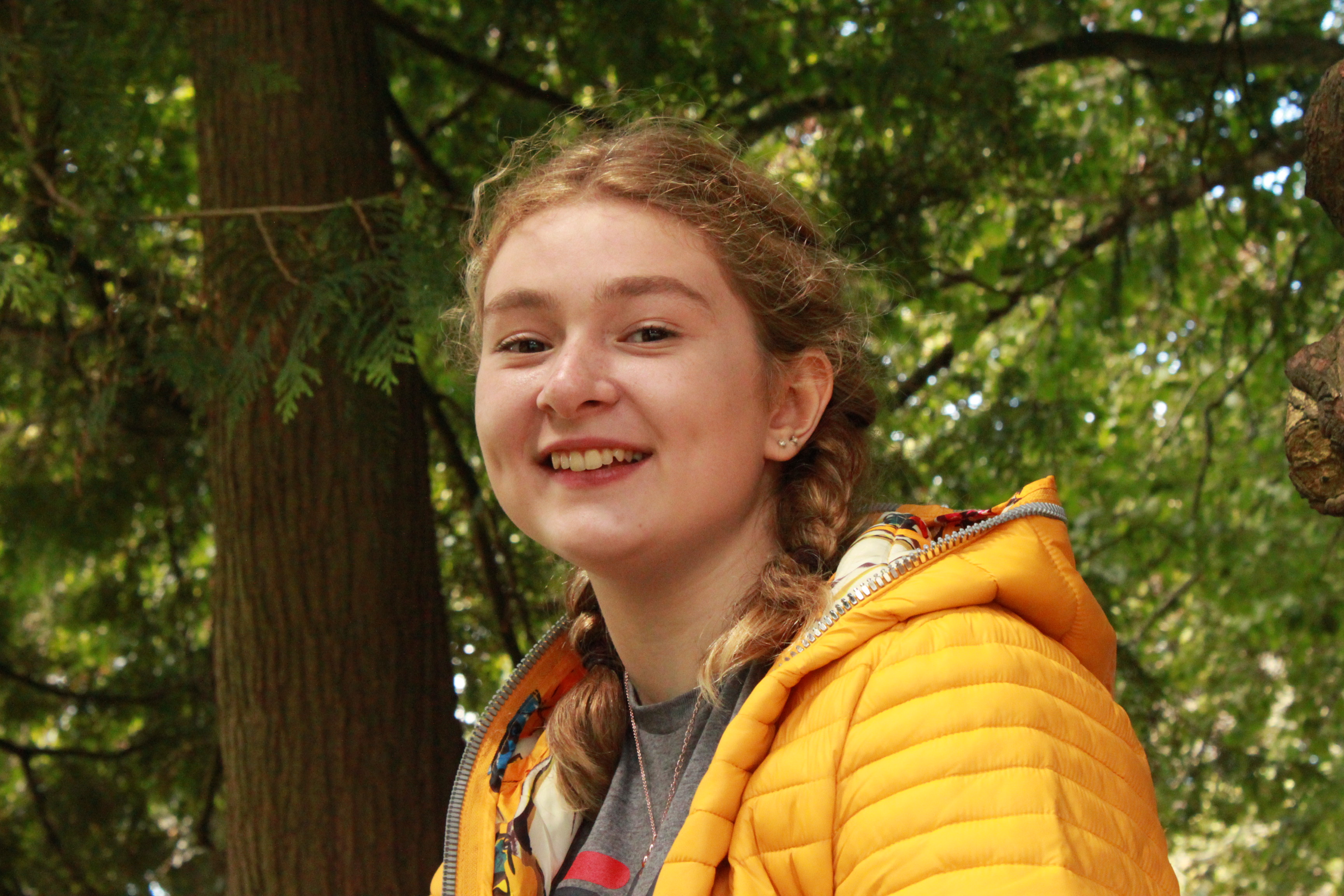 an image of a white female in a yellow coat, sat in a tree and smiling into the camera.