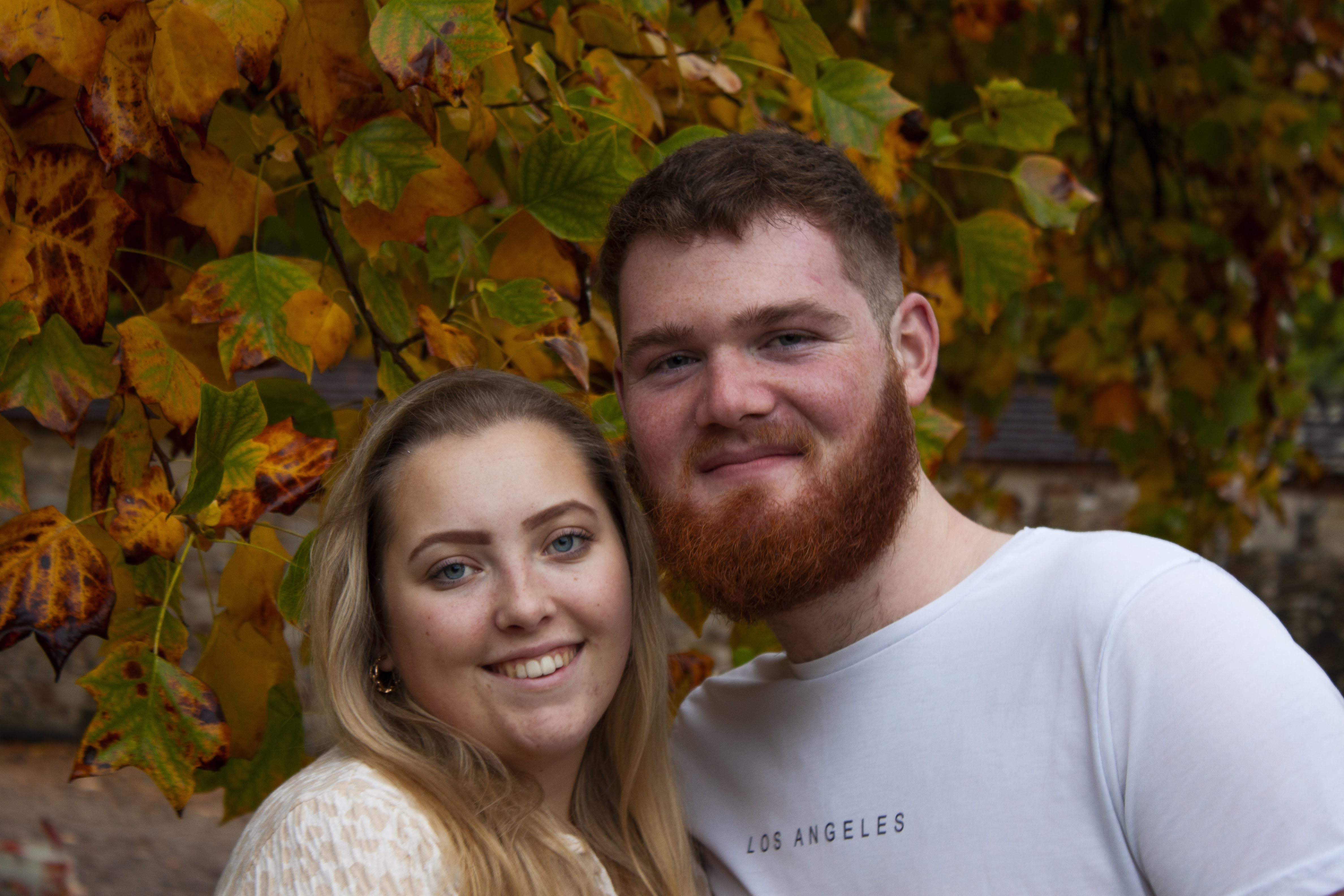 photograph of a young straight couple who are smiling into the camera with autumn leaves behind them.