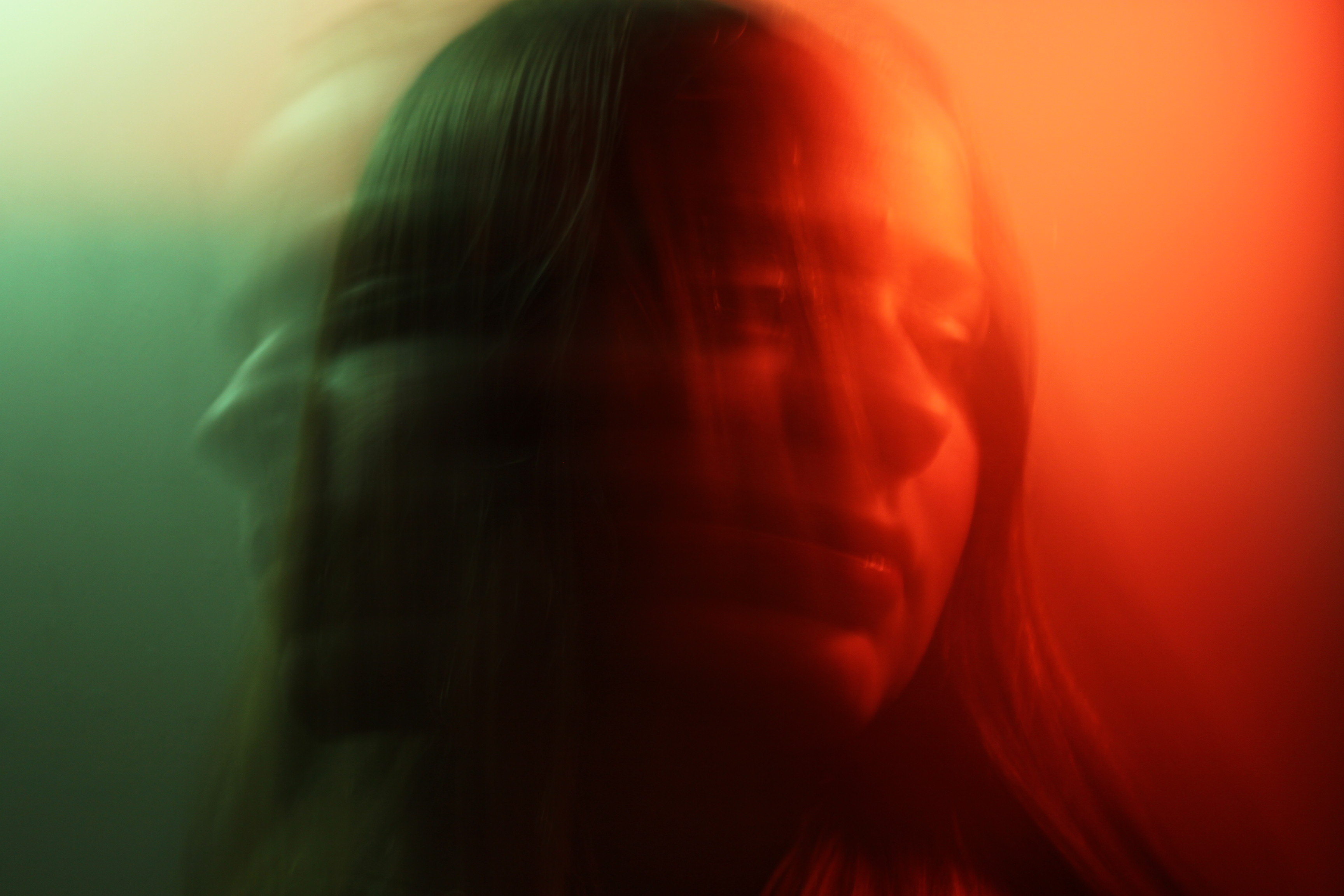 An image shot on long exposure which makes the image burry. Darcy, a white female teenager, is seen shaking her head from side to side with a red light on the left side and a blue light on the right hand side with a motion blur.